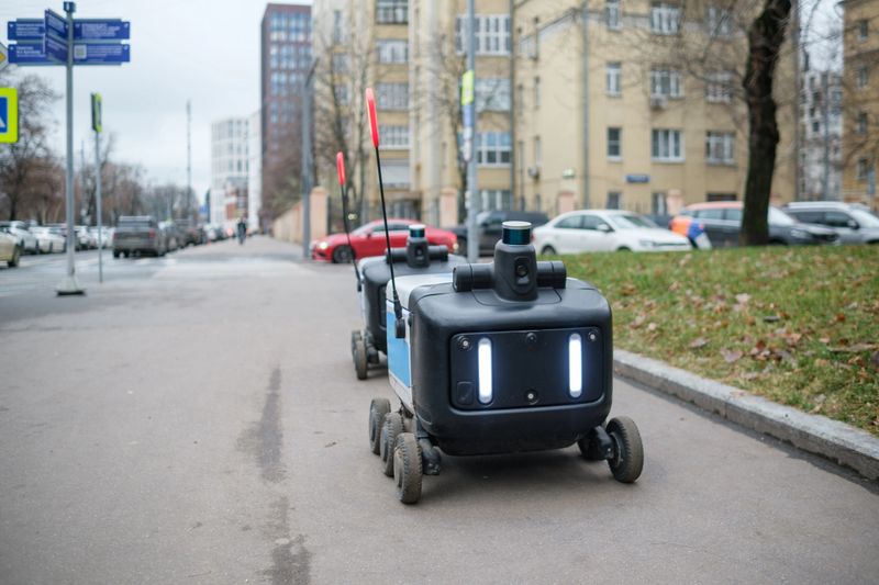 Two delivery robots navigate a city sidewalk, showcasing technology amidst urban buildings and parked cars.