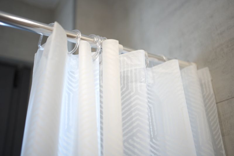 A white shower curtain hangs from a metallic rod in a sleek bathroom. The curtain features a subtle pattern, adding a touch of elegance. Soft lighting enhances the clean, modern atmosphere.