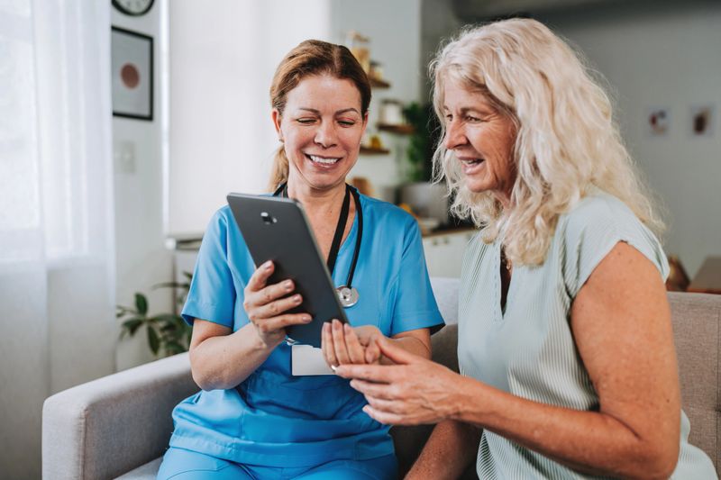 A happy healthcare professional shows medical records on a digital tablet to a senior woman during a home visit. Concept of digital health, good news, and technology