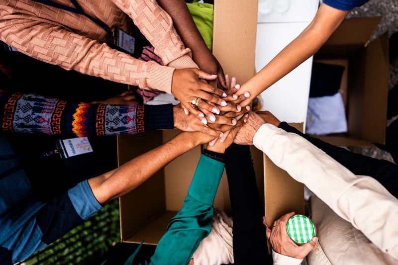 A powerful close-up image capturing diverse hands stacked together, representing unity, collaboration, and shared purpose during a community service or donation initiative. The hands showcase different skin tones and sleeve colors, symbolizing inclusivity and collective effort.