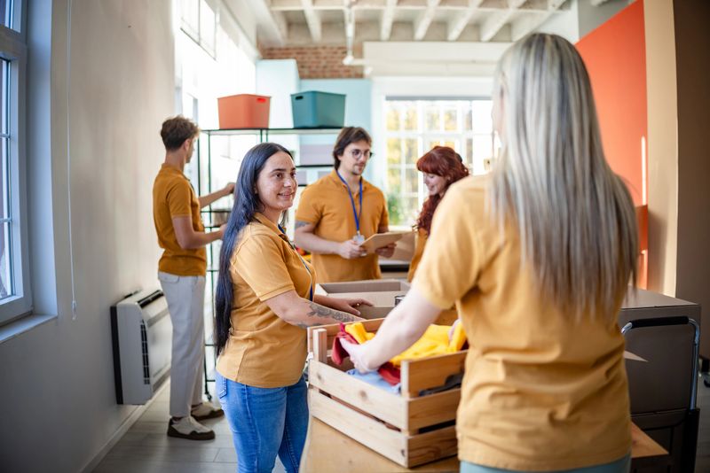 A diverse group of volunteers is working together in a food bank, smiling and arranging donation boxes filled with supplies. Their teamwork showcases generosity and community spirit.