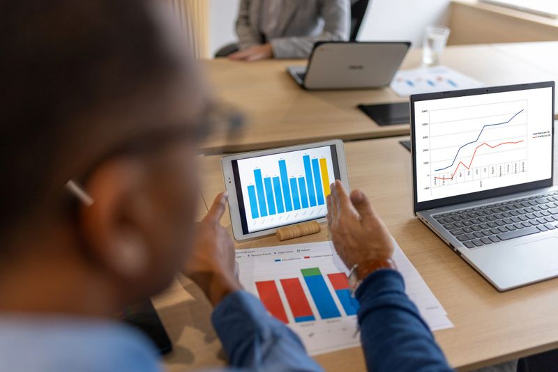 A professional compares data across a laptop and a tablet, surrounded by printed reports and coffee. Strong visual for topics including financial planning, market research, and collaborative data review.