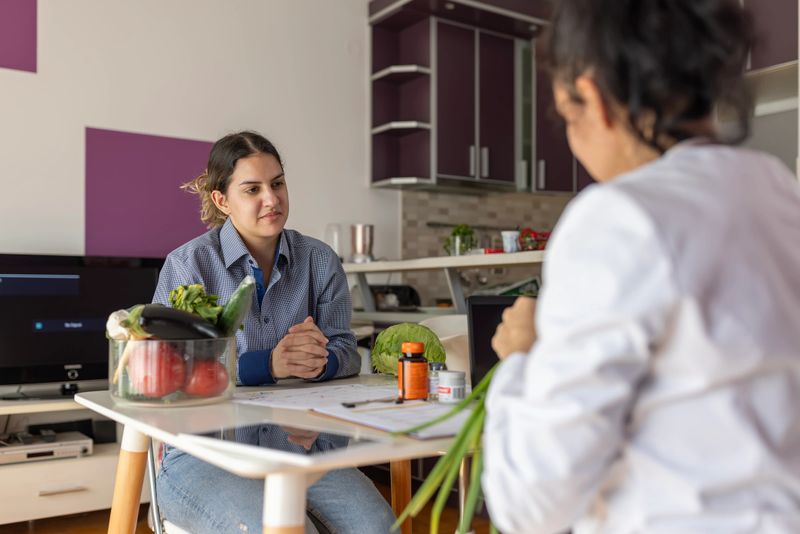 Young adult woman having a consultation with a nutritionist at home, discussing a personalized healthy eating plan with fresh vegetables visible on the table