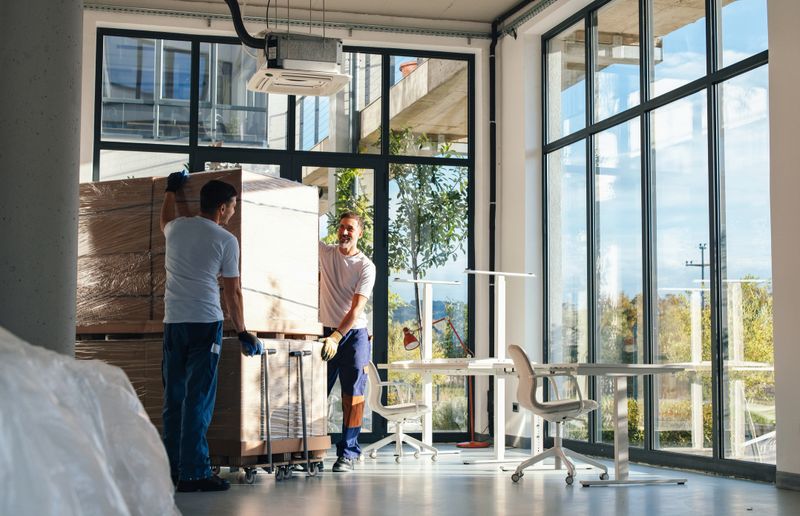 Two workers in a bright, glass-walled office lift and move large boxes on a dolly, suggesting a fresh office setup, relocation, teamwork, and logistics in a corporate environment.