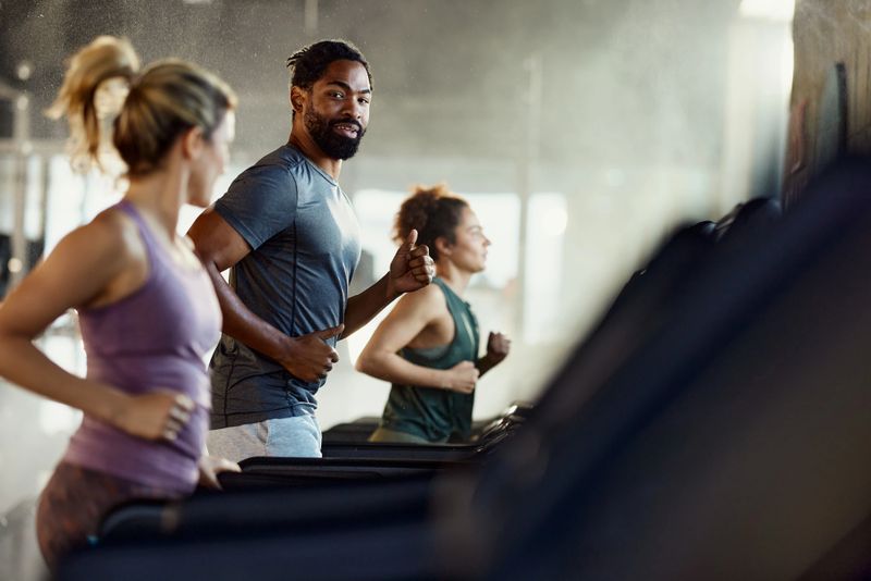 Group of athletes running on treadmills during sports training in a health club. Focus is on black man talking to his friend.