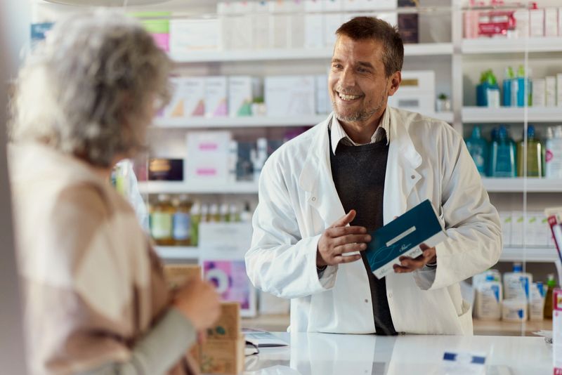 Happy male pharmacist communicating with his customer about nutritional supplements in a pharmacy. The view is through glass.