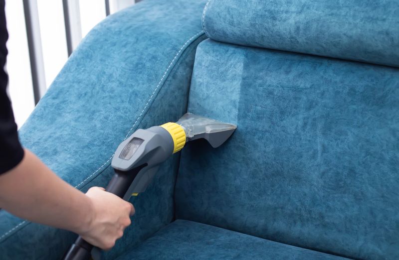 A person is using a vacuum cleaner to clean a blue sofa in a well-lit living room. The cleaning process focuses on the seams and crevices of the furniture.