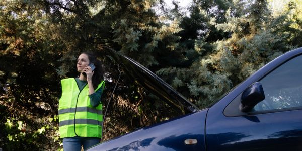 Woman in safety vest calls for help beside a broken-down car with hood open.