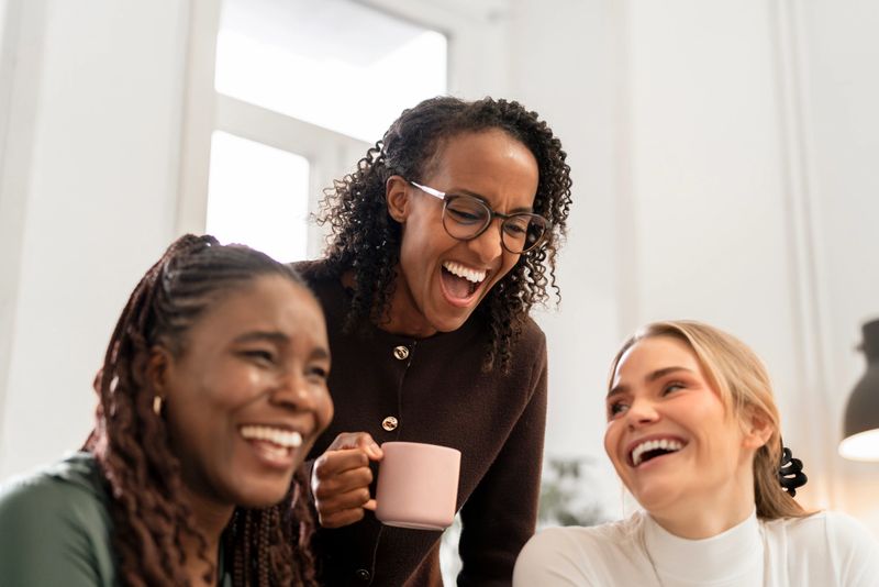 Three diverse women share a joyful moment, laughing and chatting in a sunny, modern space. One holds a pink coffee mug, highlighting friendship, warmth, and positive energy in a casual gathering. Soft focus.