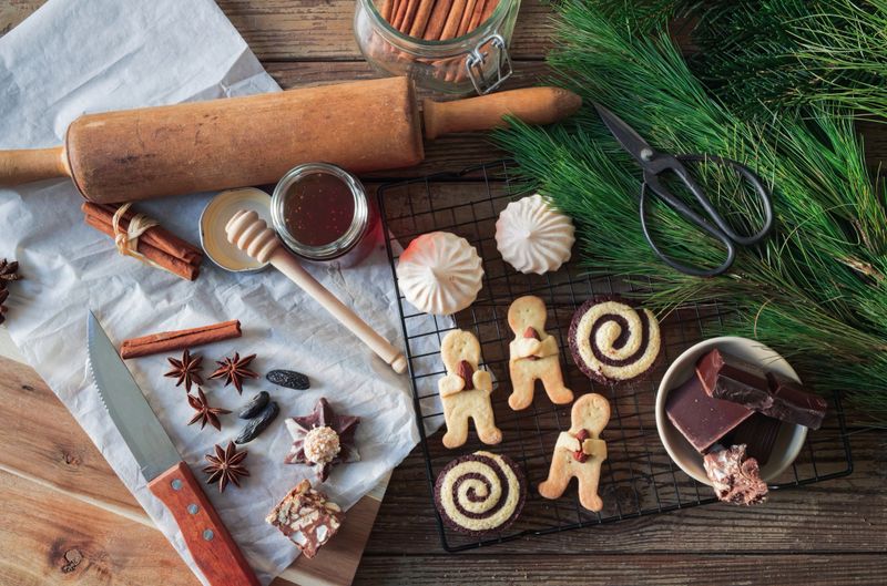A cozy top-down view of homemade Christmas cookies, chocolate, star anise, cinnamon sticks, and a honey jar arranged on a rustic wooden table. A rolling pin, scissors, and fresh pine branches add a festive and natural touch. Ideal for eco-conscious holiday baking, traditional recipes, and slow-living seasonal visuals. Copy space on parchment and wooden surface allows for custom holiday messaging.