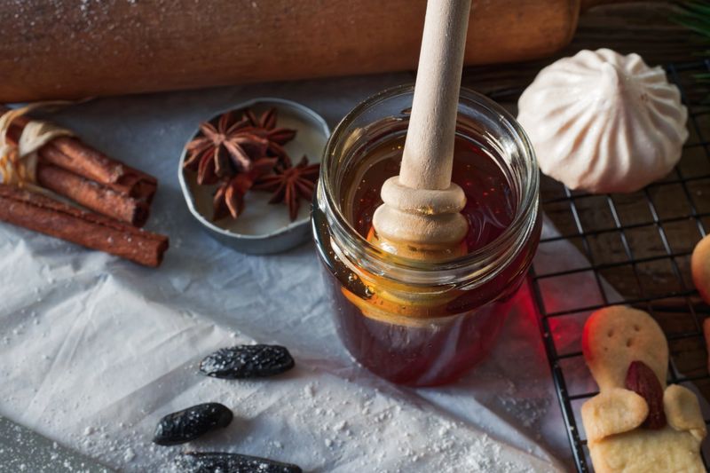 A close-up of a glass honey jar with wooden dipper surrounded by festive holiday cookies and winter spices. Cinnamon sticks, star anise, and a single meringue create a comforting, natural vibe. Perfect for sustainable holiday cooking visuals. Copy space included for seasonal messages or recipe text.