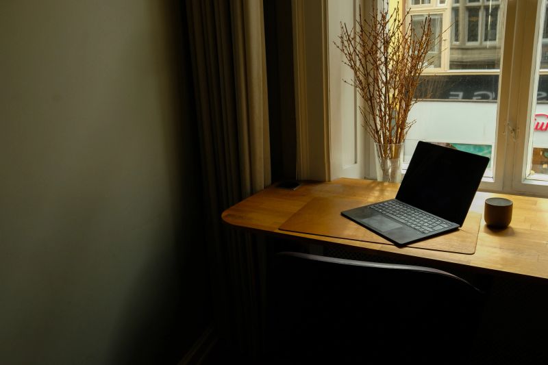 A clean and minimalist home office workspace featuring an open laptop, a ceramic mug, and a vase of dried branches placed on a wooden desk beside a large sunlit window. This warm, modern scene represents remote work, digital productivity, flexible work culture, and contemporary workspace design. Ideal for concepts such as freelancing, creative work, hybrid work environments, focus time, and digital nomad lifestyle.