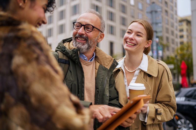 Three colleagues stand together in a modern urban area, smiling and discussing information displayed on a tablet. One man holds the device while the two women beside him carry coffee and documents, creating a relaxed, collaborative business moment outdoors.