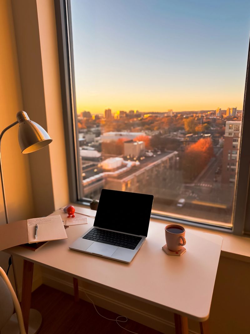 Cozy home workspace with laptop, notebook and mug on a desk by a large window, overlooking a city skyline at sunset.