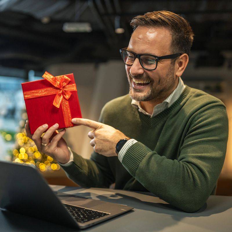 Man smiling and holding a red gift box while engaging in a video call on a laptop, celebrating a special occasion with a christmas tree in the background