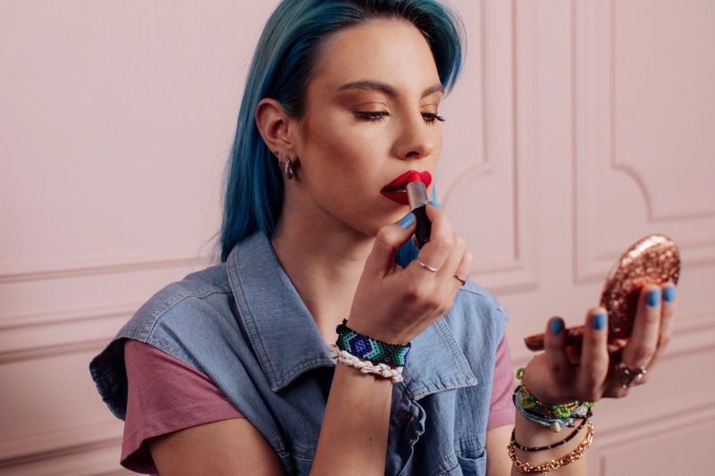 A fashionable woman with vibrant blue hair applies bold red lipstick in a chic, pink-toned studio; She wears a denim vest and colorful bracelets, exuding modern beauty and self-expression