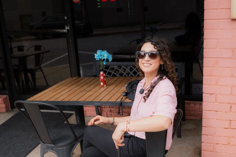 Cheerful woman wearing sunglasses and a pink shirt sitting at an outdoor cafe table with a vase of blue flowers