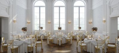 Elegant white and gold dining room with round tables and tall arched windows.