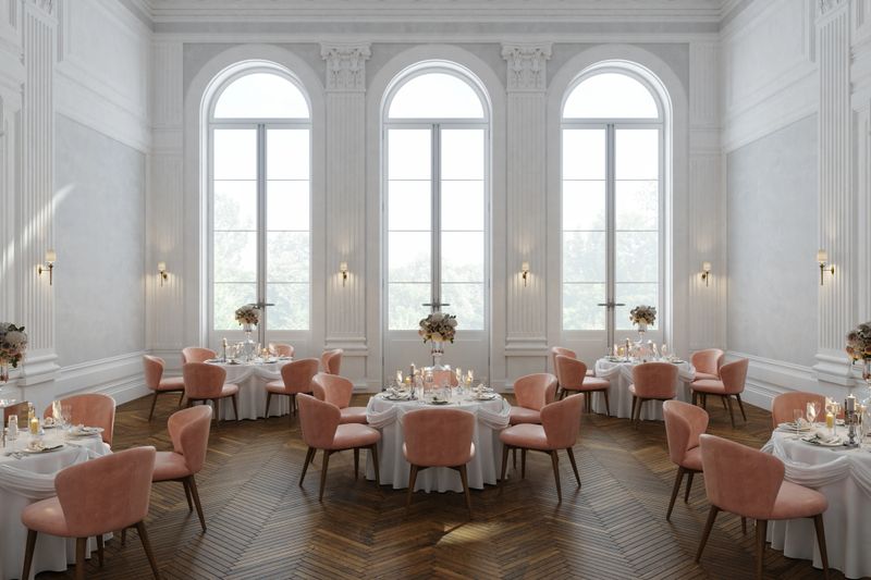 A bright and elegant banquet hall featuring round tables, soft pink chairs, floral centerpieces, and large arched windows creating a refined and romantic atmosphere.