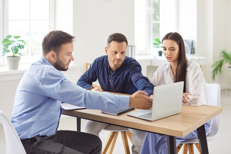 Young family couple sitting at desk with real estate agent, looking at laptop screen, choosing new house. Husband and wife exploring real estate options for purchase or rent with professional real estate agent.