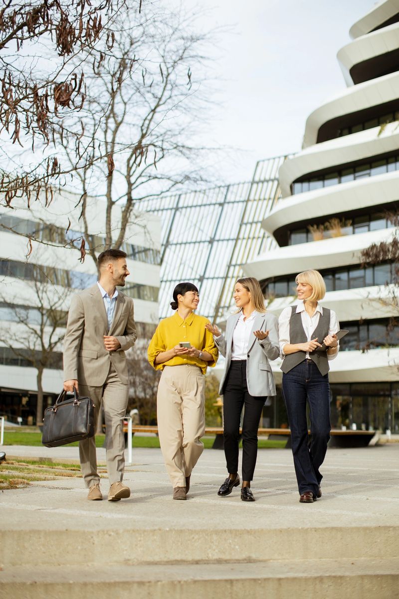 Four business professionals are walking together on a sunny day in a modern urban area. They appear engaged in conversation, showcasing collaboration and teamwork amid contemporary architecture.