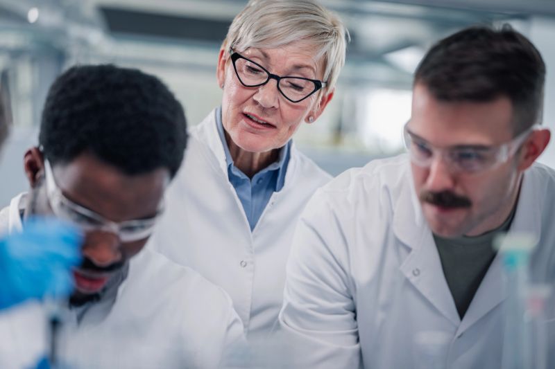 Diverse group of scientists in lab coats and safety glasses discussing progress and conducting experiments in a bright, modern research facility