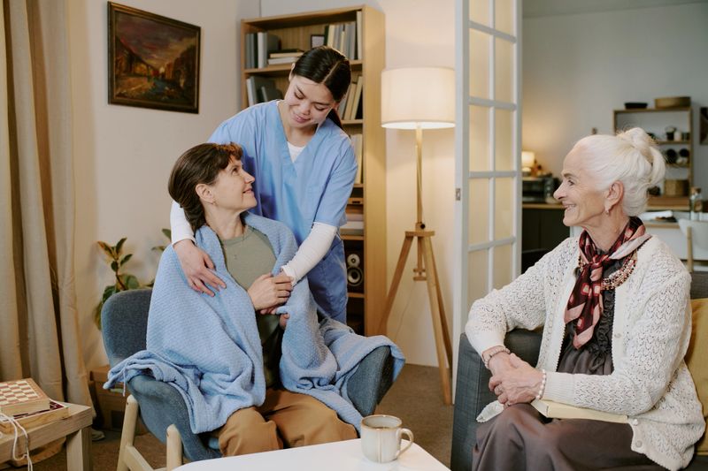 Middle aged Caucasian woman sitting in armchair covered with blanket being comforted by young adult Hispanic female nurse, while senior Caucasian woman sitting nearby smiling