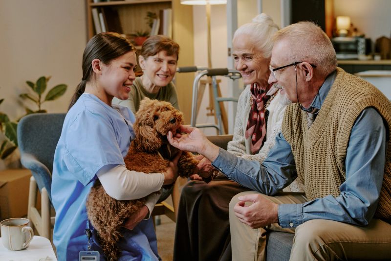 Group of senior Caucasian women and senior Caucasian man interacting with young adult Caucasian female caregiver holding small dog, all smiling and engaging in friendly conversation