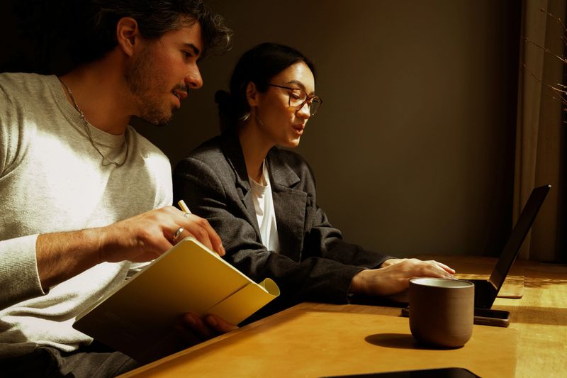 Two coworkers sit together at a laptop, having a collaborative meeting and brainstorming session beside a large window filled with natural light. The bright, modern workspace creates an atmosphere of creative thinking, teamwork, and productive problem-solving. Their focused interaction captures the energy of contemporary office culture, remote collaboration, and innovative business environments. This scene reflects modern coworking, digital workflow, and professional communication, making it ideal for themes related to project planning, startup teamwork, and business strategy development.