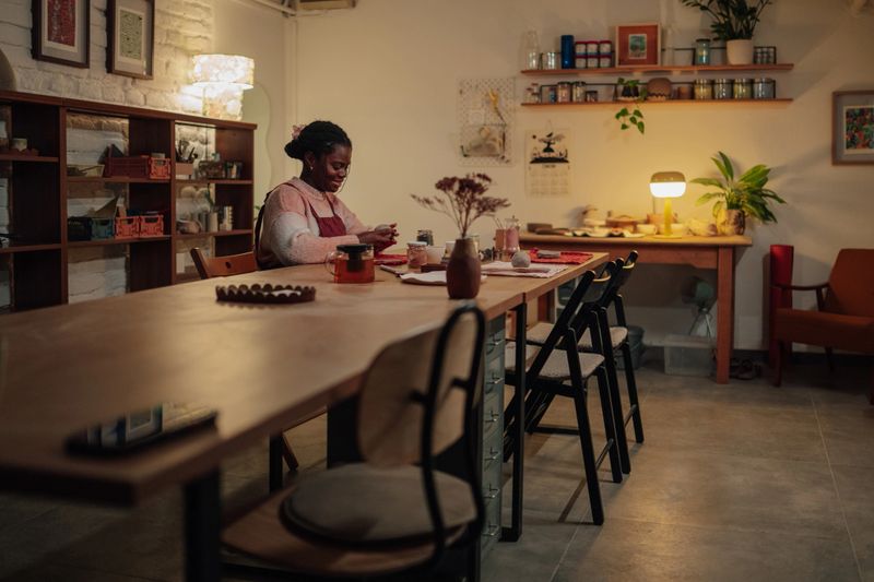 Young adult woman smiling, enjoying a cup of tea while doing a creative craft activity at a cozy home workshop in the evening