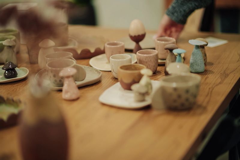 Artisan displaying unique pottery, including small cups and decorative mushroom figures, on a rustic wooden table, highlighting skilled craftsmanship