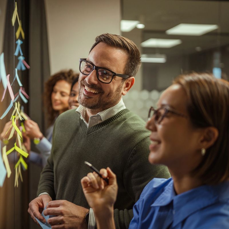 Group of diverse business professionals collaborates during an office meeting, brainstorming innovative ideas and posting colorful sticky notes on a clear glass whiteboard in a modern workspace