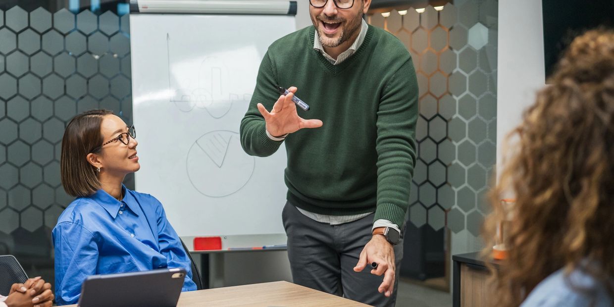 Man in green sweater enthusiastically presents to colleagues in a modern office.
