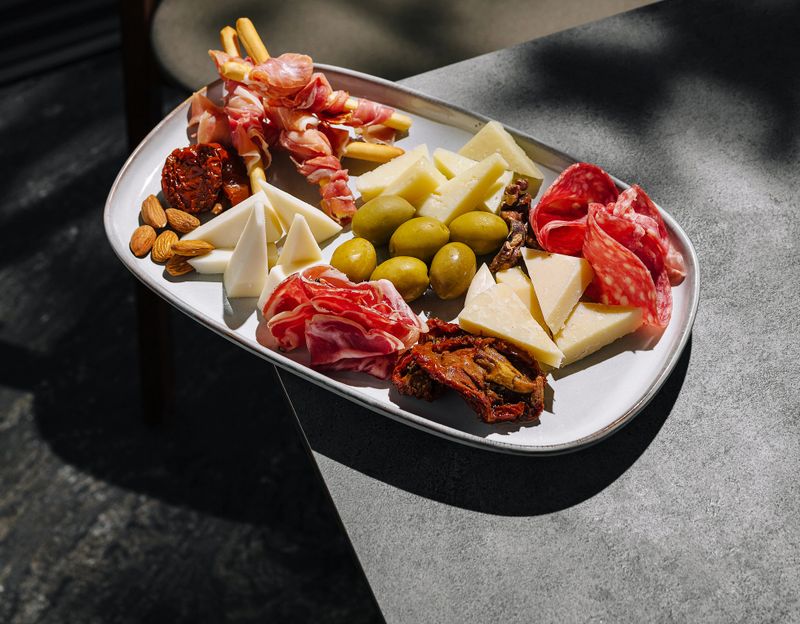 A carefully arranged charcuterie board features various meats, cheeses, olives, and nuts. Sunlight casts shadows on the table, enhancing the outdoor dining experience.