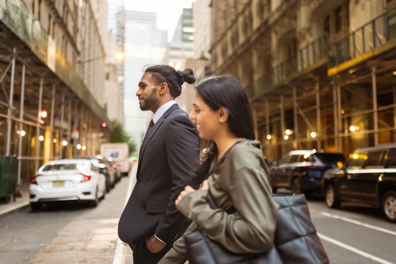 A man in a suit and a woman wait to cross a city street surrounded by tall buildings and cars during the day.