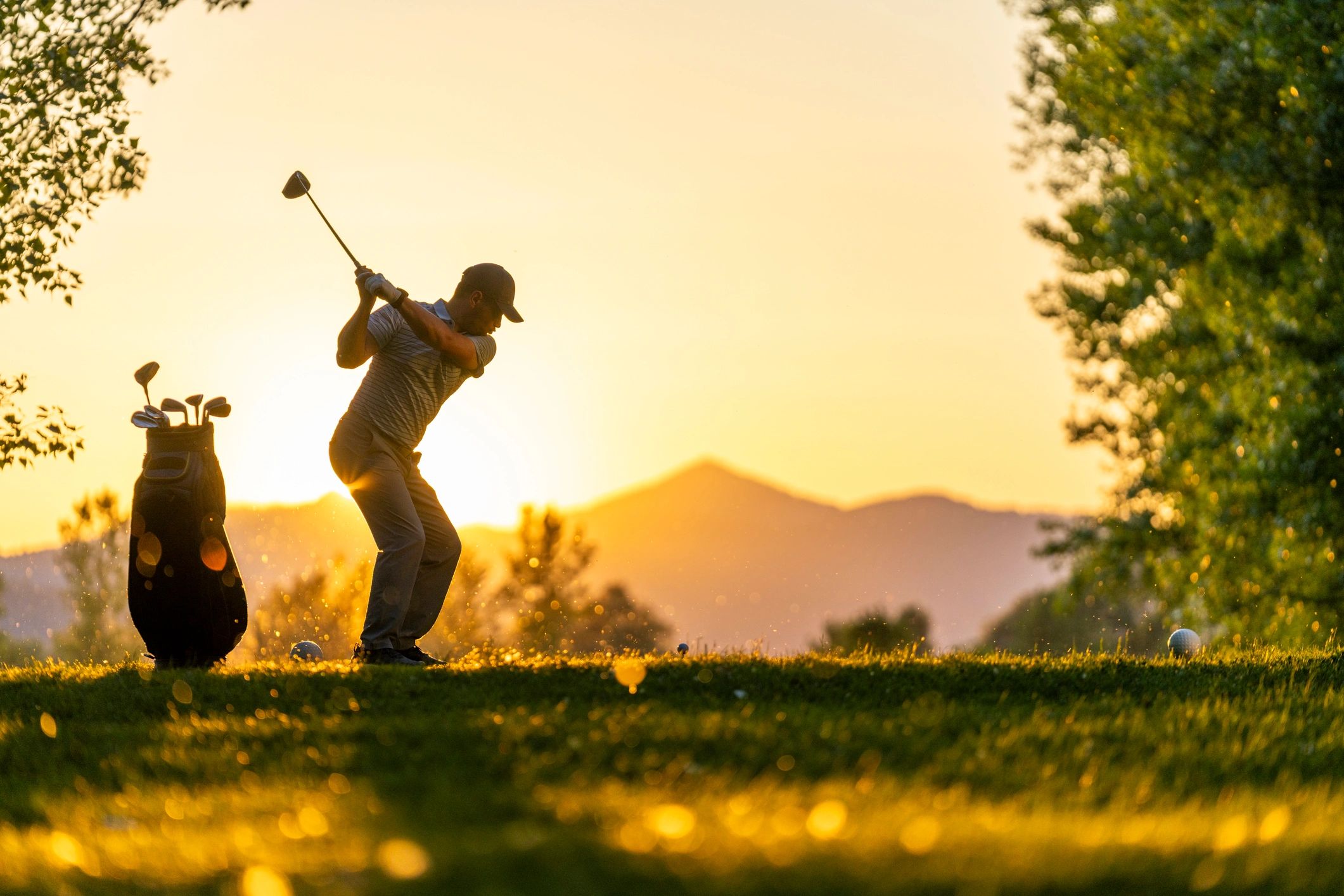 Golfer swinging club at sunset with bag of clubs nearby.