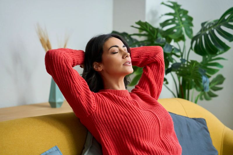 Woman in red sweater relaxing on yellow sofa with hands behind head, enjoying quiet moment