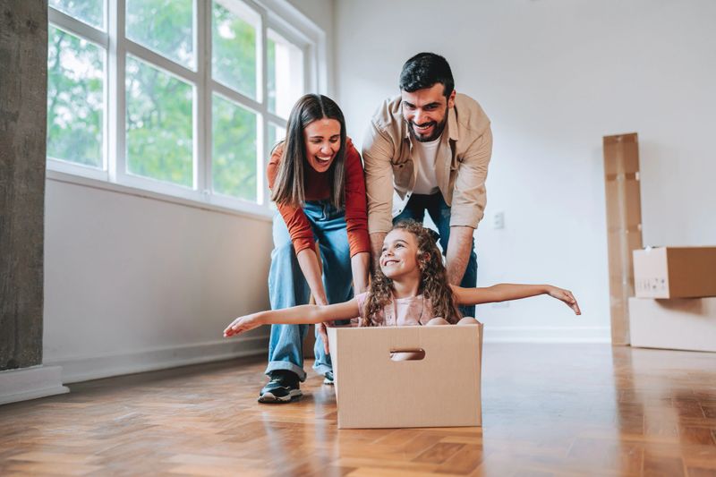 Smiling parents playing with their daughter sitting inside a cardboard box in an empty apartment during moving day. Concept of family fun, new beginnings, home ownership, and happiness in the new house