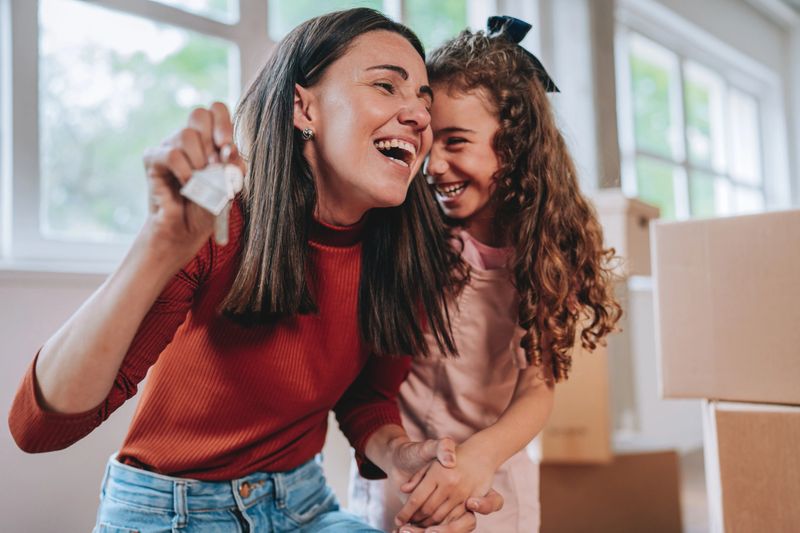 Happy mother holding house keys and smiling at her daughter inside an empty apartment with moving boxes. Concept of love, family connection, new home ownership, and life achievements