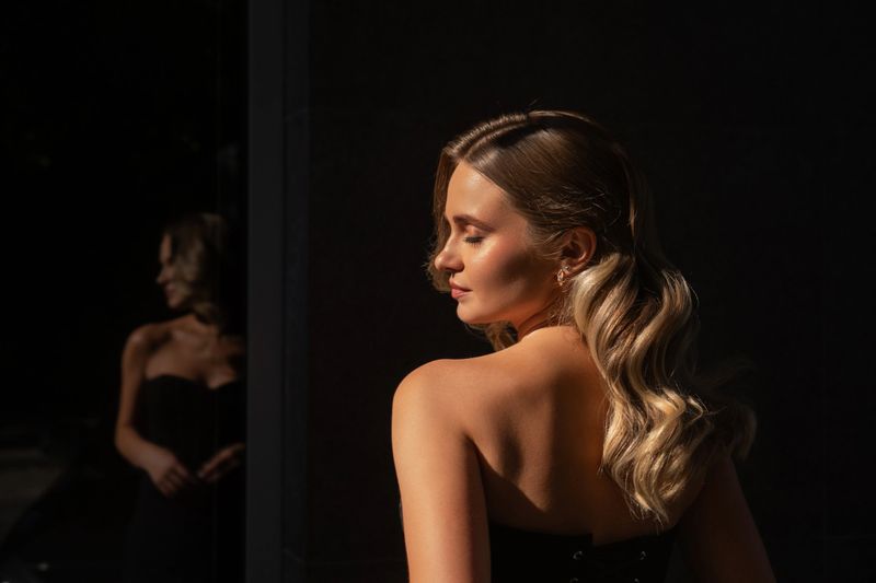 A model displays a serene expression while posing in soft sunlight. Her long hair cascades down her back, creating an elegant contrast against the dark setting.