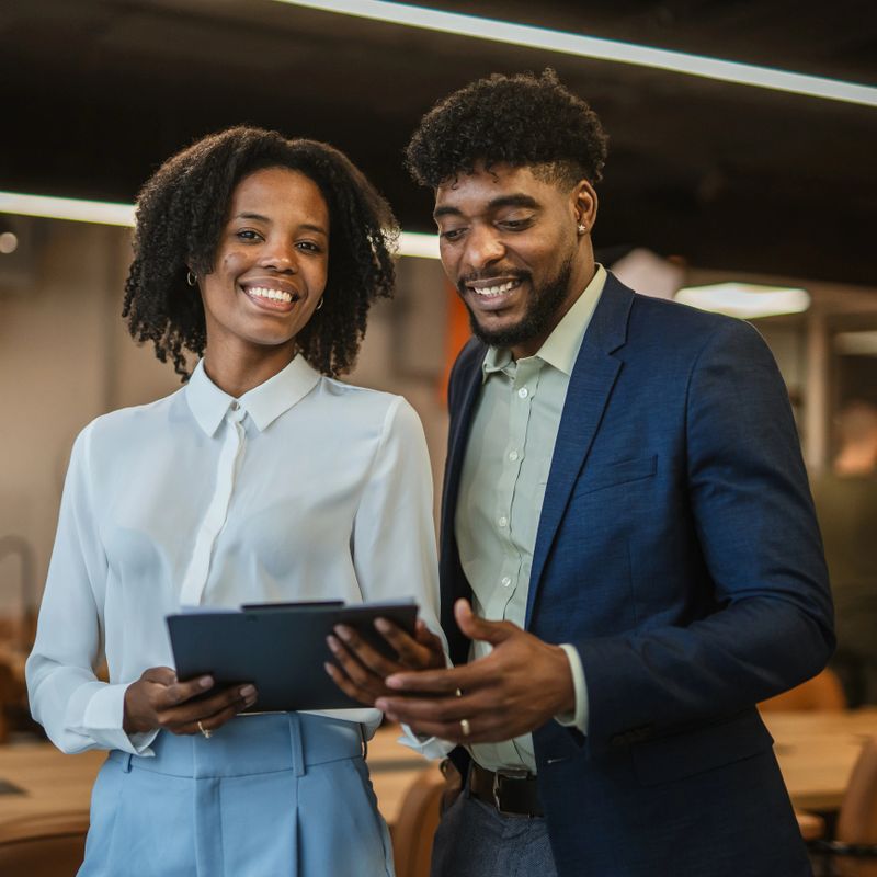 Two professional african american business colleagues smiling and collaborating while discussing work on a digital tablet in a bright, modern office environment
