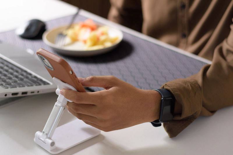 close-up shot focused on a male hand and wrist wearing a black digital smartwatch, interacting with the touch screen of a smartphone propped on an white desk stand. Concepts of technology, remote productivity, and personal wellness/fitness tracking