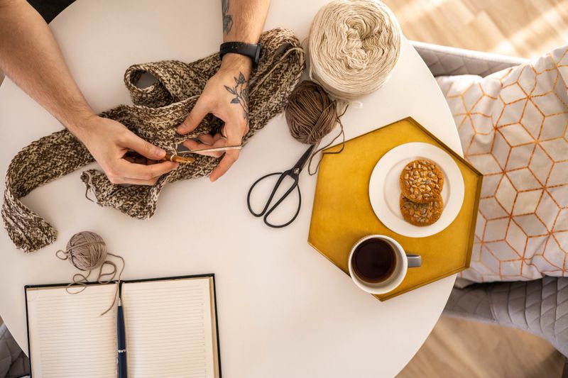 Feminine male hands crochet handmade artwork hobby on wooden table with sewing tools closeup top view. Man arm art creative hobby use hook and wool yarn handmade warm clothes and accessories workshop