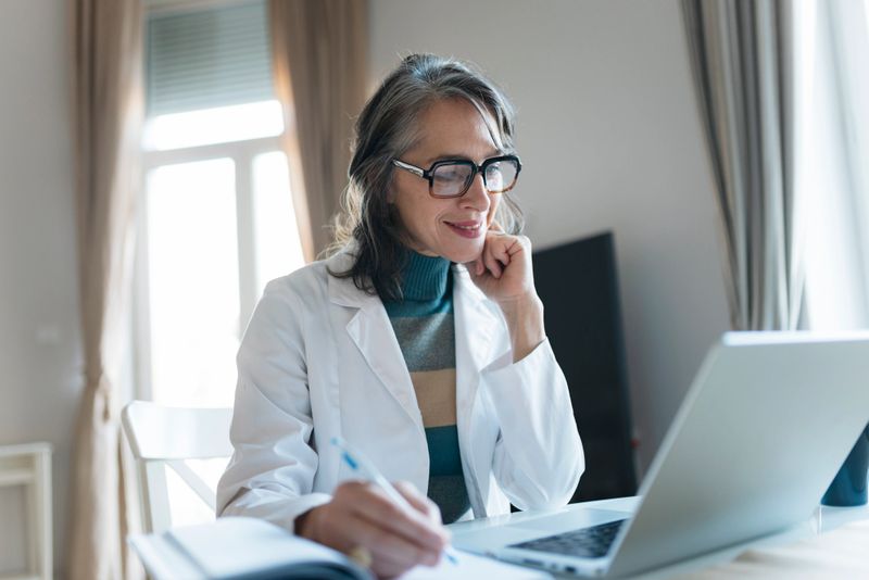 Mature female doctor in lab coat teleworking, smiling while looking at laptop and writing