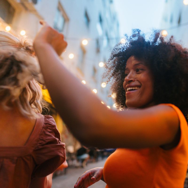 Photo of two cheerful female friends, dancing on the street under the string lights