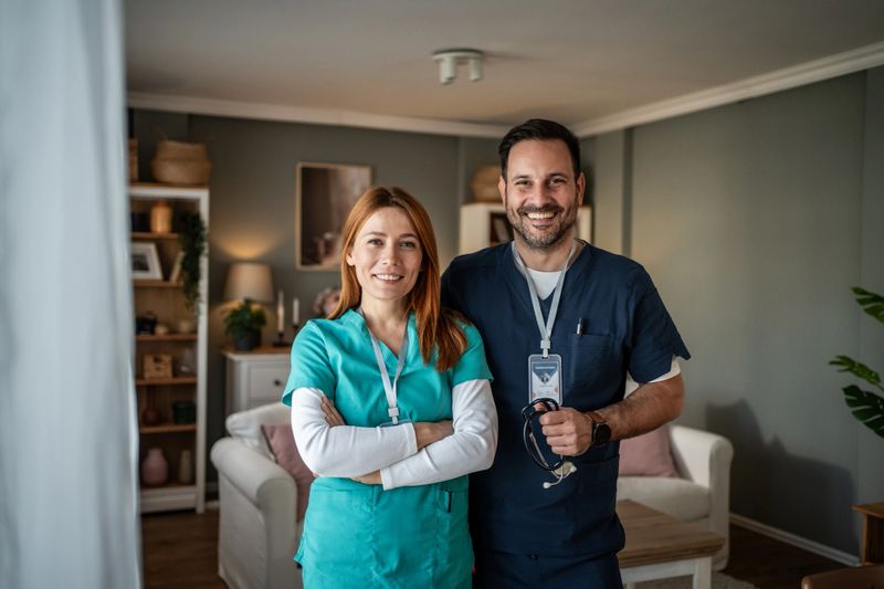 Smiling male and female healthcare workers wearing scrubs and id badges, standing in a residential living room, reflecting concepts of teamwork, compassionate care, and medical house calls