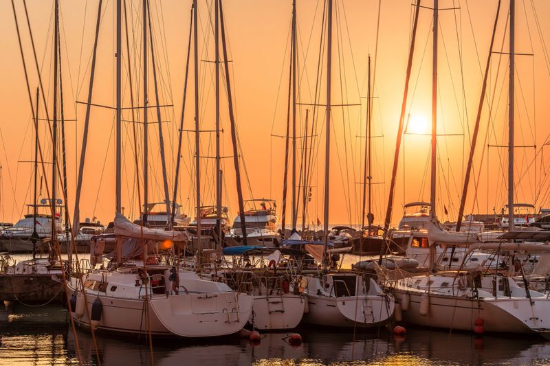 An orange sunset over the sea, with a view of boats moored at the shore. The sun is low on the horizon behind the masts of sailboats, and the seaside landscape is visible from the shore.