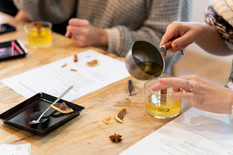 Woman's hands pouring essential oil infused soy wax into a glass jar with a wooden wick during a candle making workshop