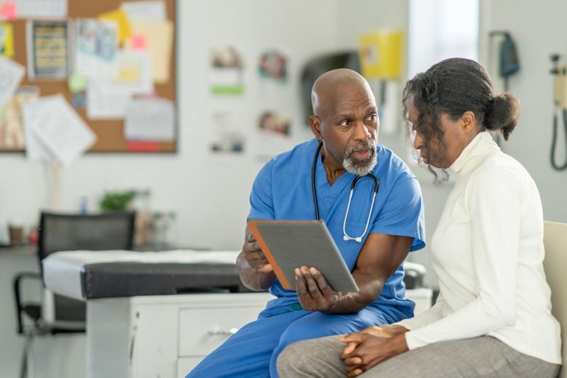 A compassionate Black male doctor in blue scrubs sits with an older Black woman in a clinical office, reviewing notes on a tablet. They discuss health, care, and medical advice, conveying trust, guidance, and patient-centered care.