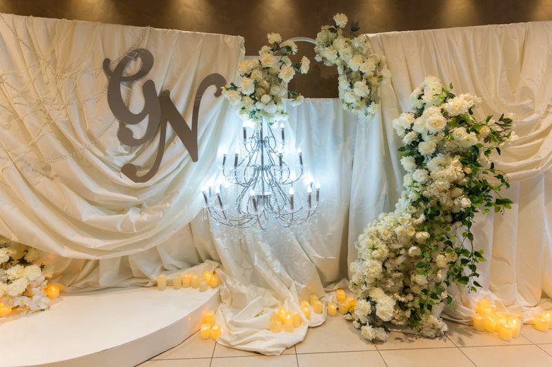 White drapes hang in an event space while a chandelier is illuminated. Floral arrangements are present, creating a festive atmosphere for a wedding celebration.